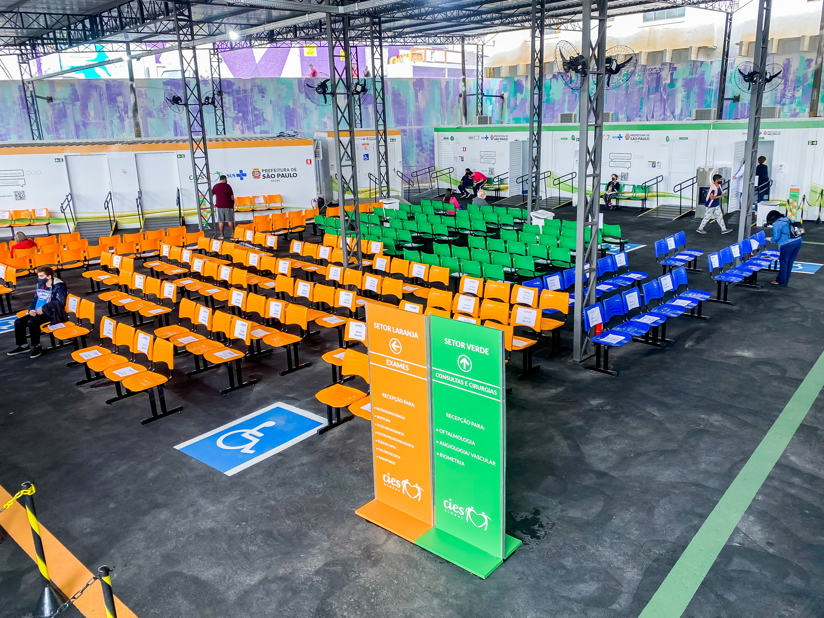 Foto de espaço - Cercada por contêineres brancos, uma ampla sala de espera se forma com cadeiras de cores amarelas, verdes e azuis, Há totens de sinalização e marcações no chão para cadeirantes